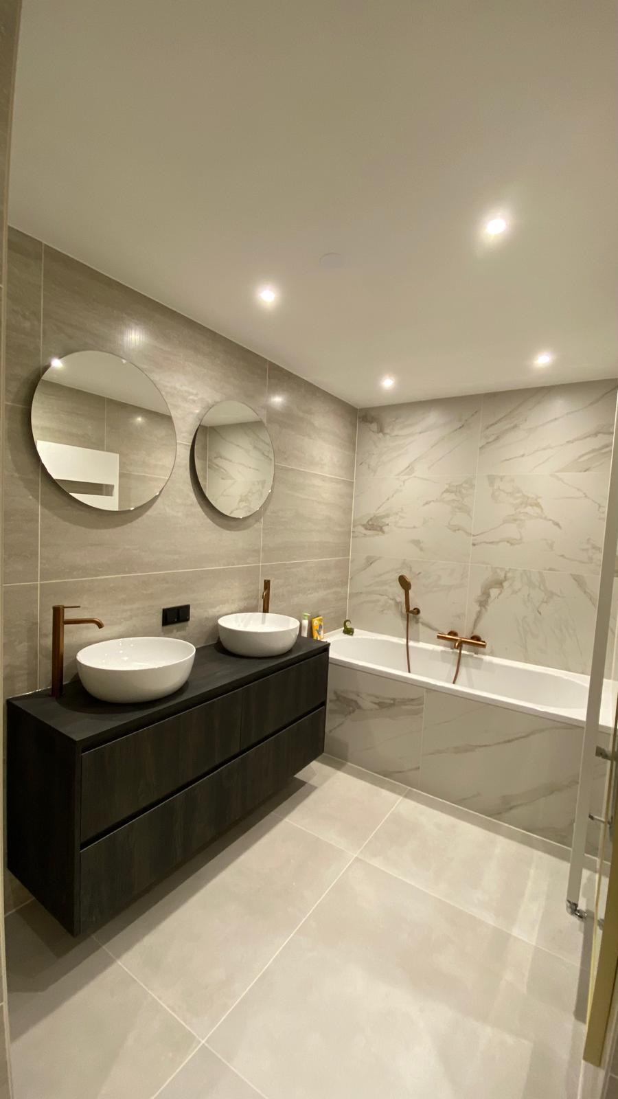 Ruime badkamer met ligbad, dubbele wastafel, ronde spiegels en goudkleurige kranen.