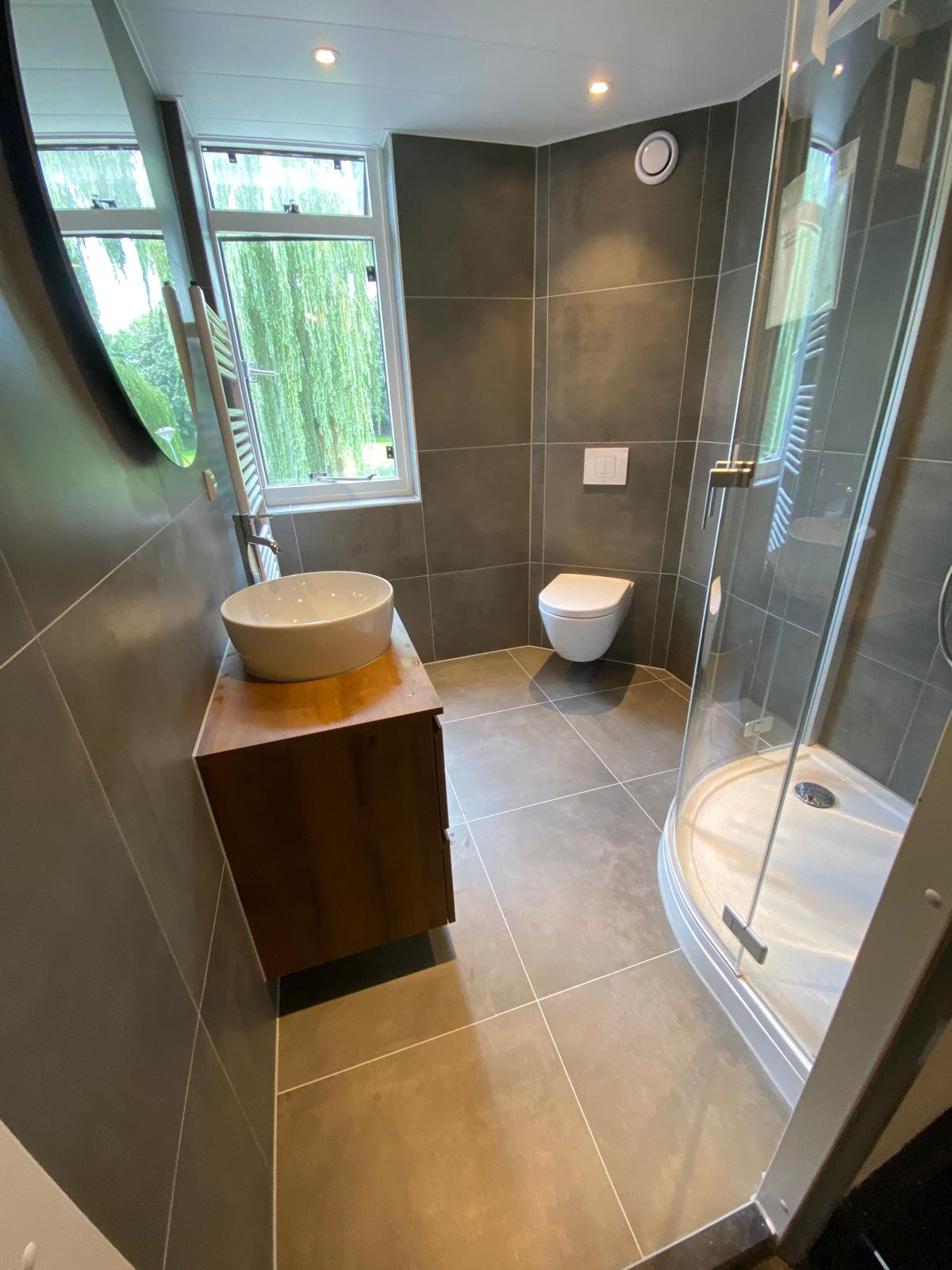 Badkamer met donkere wandtegels, hoekdouche, wandtoilet en houten wastafelmeubel.