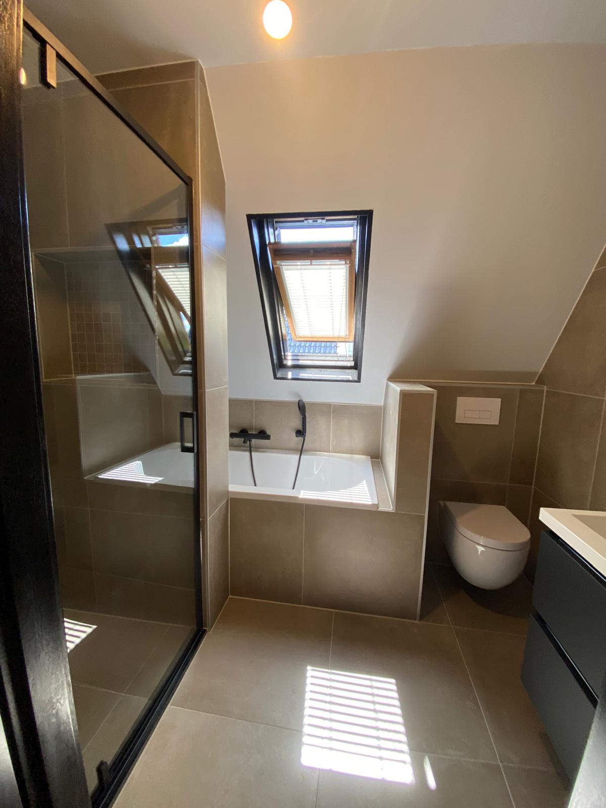 Badkamer onder een schuin dak met ligbad, inloopdouche en donker wastafelmeubel.