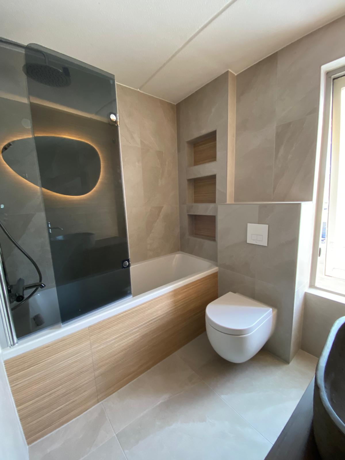 Badkamer met ligbad, glazen douchescherm, wandtoilet en ingebouwde nissen.
