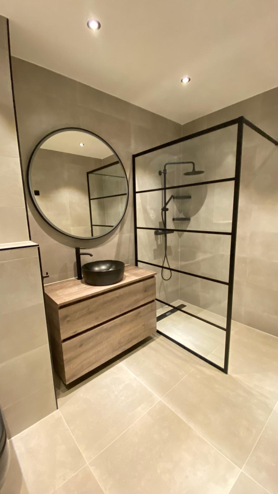 Badkamer met houten wastafelmeubel, ronde spiegel en zwarte glazen douchewand.