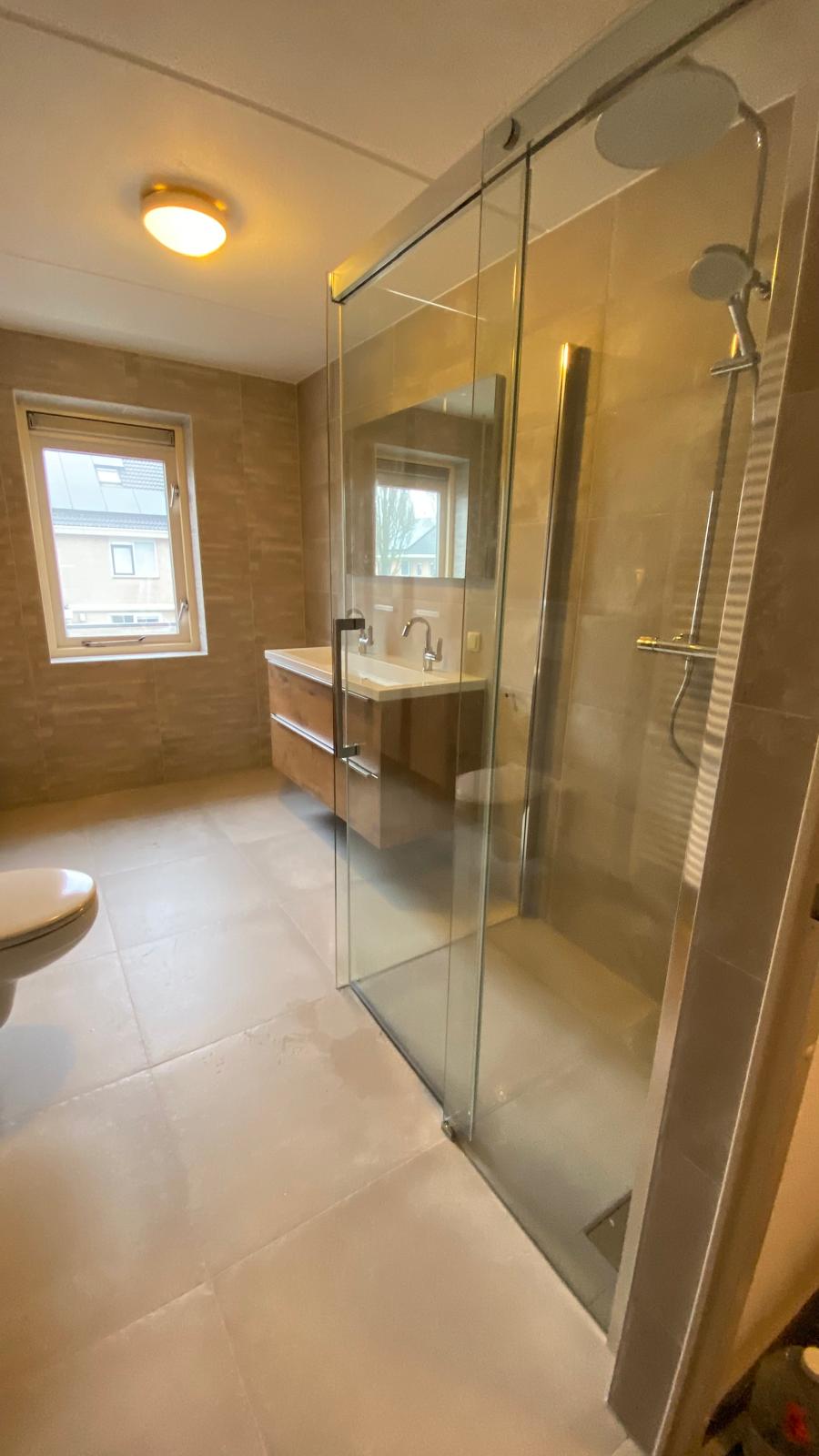 Afgewerkte badkamer met glazen douchewand, wastafelmeubel en grote grijze tegels.