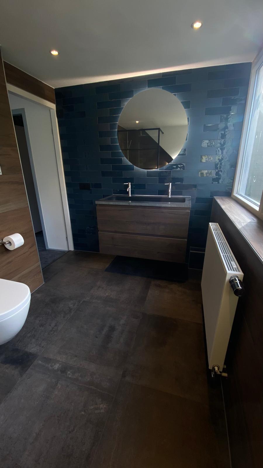 Badkamer met donkerblauwe wandtegels, een houten wastafelmeubel en ronde spiegel.