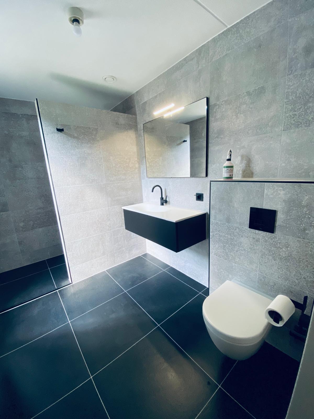 Ruime badkamer met inloopdouche, wandtoilet, donkere vloer en zwart kraanwerk.