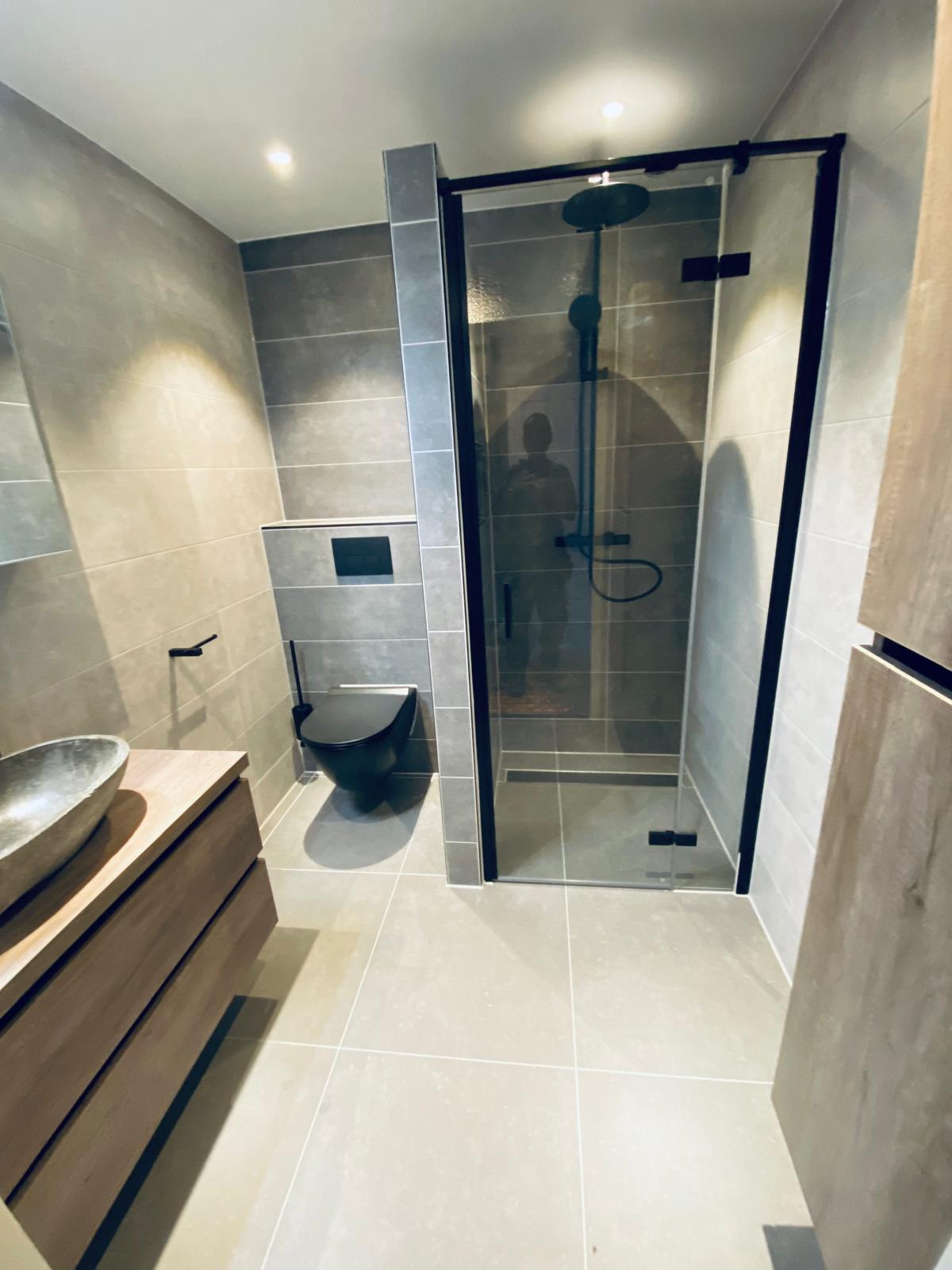 Badkamer met houten wastafelmeubel, wandtoilet en inloopdouche met zwarte accenten.