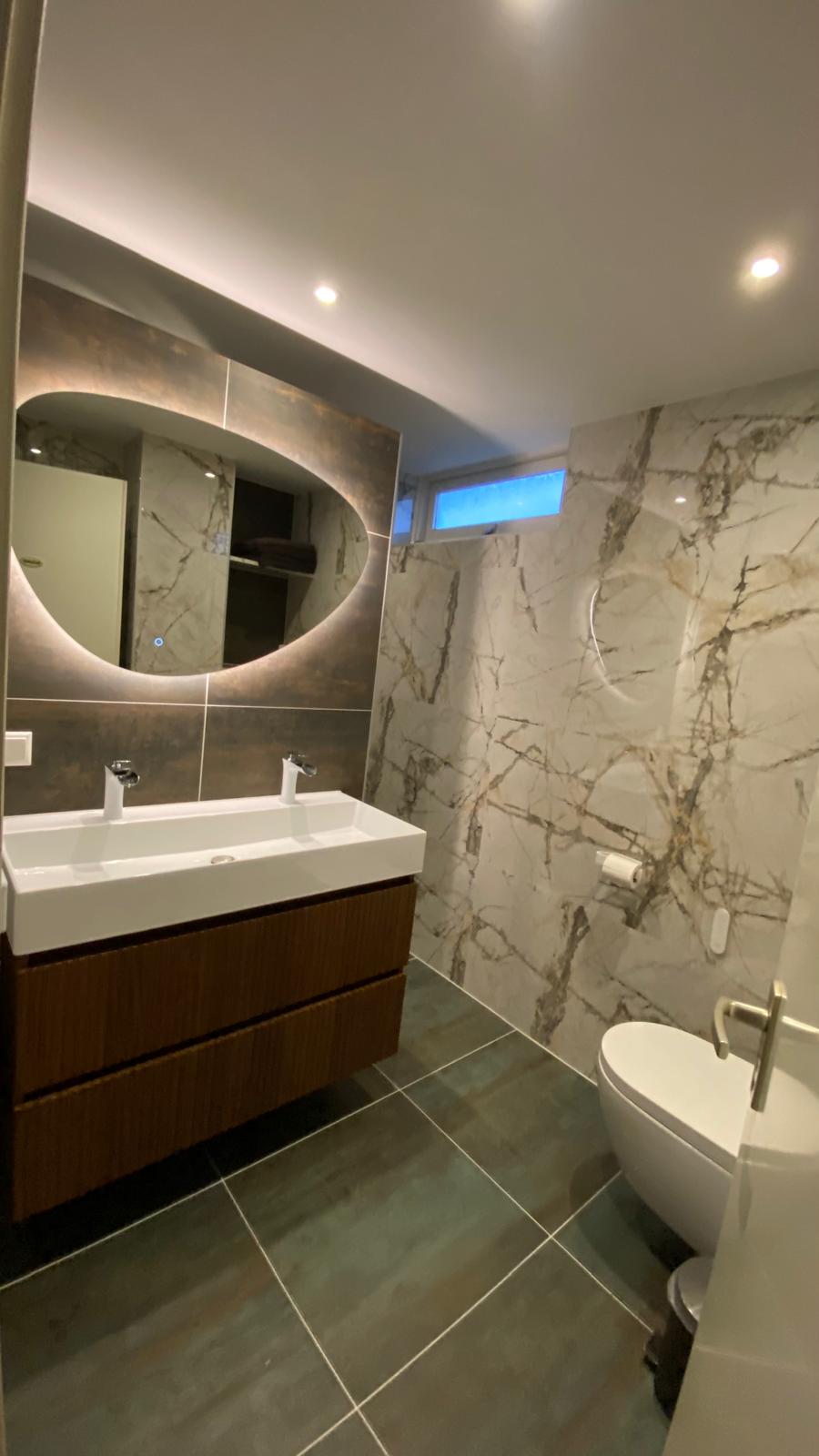 Badkamer met houten wastafelmeubel, verlichte spiegel en marmerlook douchewand.